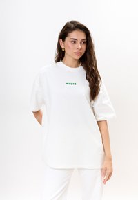 Vrouw met lang bruin haar, die een witte oversized T-shirt draagt met "MIRONS" in groene tekst, witte broek, gouden oorringen en een parelketting.