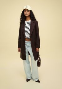 Brown oversized coat, white and navy striped crop top, light blue wide-leg jeans, black pointed shoes, and a yellow cap with text.