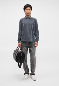 Πόλο μπλούζα μακρυμάνικη σκούρου γκρι χρώματος, ανοιχτού γκρι τζιν και λευκά αθλητικά παπούτσια, συνοδευόμενα από μια μαύρη τσάντα duffel με καφέ λεπτομέρειες. Μινιμαλιστικός σχεδιασμός και χαλαρή εφαρμογή.