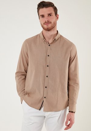 Camisa de manga larga marrón claro con collar abotonado, botones negros y una tela ligeramente texturizada, acompañada de pantalones blancos.