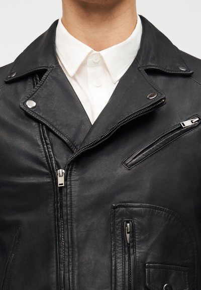 Άνδρας που φοράει μαύρο δερμάτινο μπουφάν με ασημένια φερμουάρ και κουμπιά πάνω από ένα λευκό πουκάμισο με γιακά, εμφανίζονται ο κορμός και ο λαιμός του.