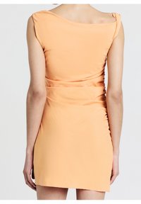 Femme portant une mini-robe sans manches couleur pêche avec de subtils fronces sur le côté droit, vue de dos sur un fond uni clair.