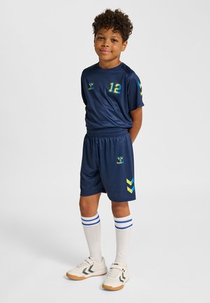 Dreng stående med hænderne bag ryggen iført navyblåt sportstøj med nummer 12, hvide knæstrømper med blå striber og hvide sneakers.