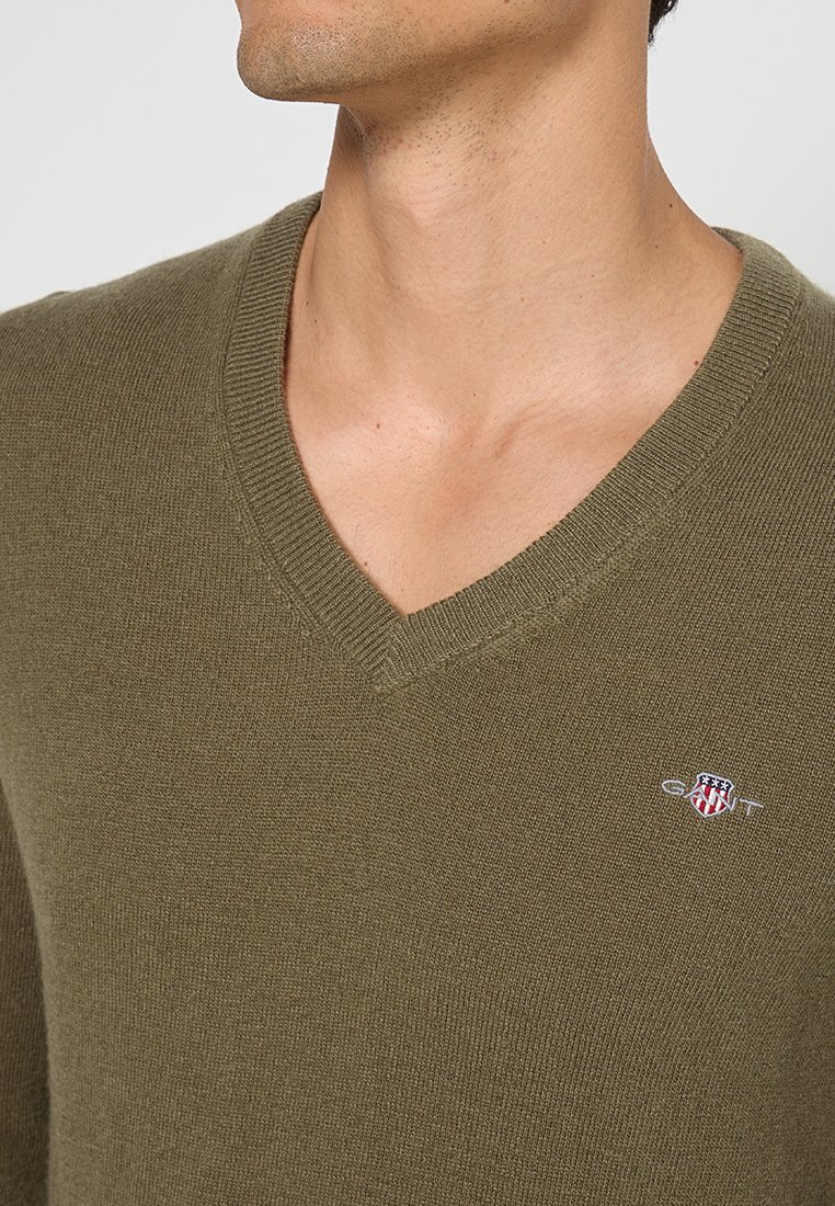 Pull à col en V couleur vert olive fabriqué en matière tricot douce. Il présente un petit logo brodé sur la poitrine. La texture est lisse et uniforme.
