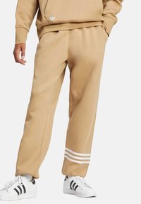 Tan sweatpants feature a loose fit, smooth texture, and three white stripes at the bottom. Paired with white sneakers with black accents.