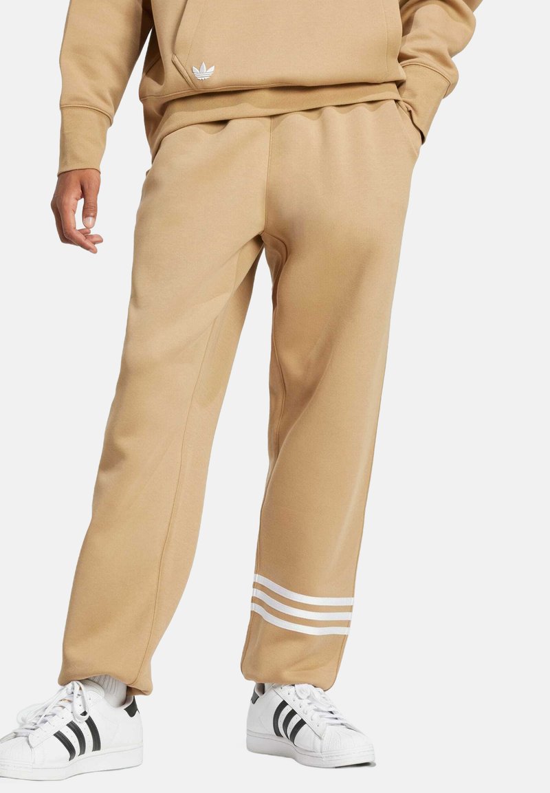 Tan sweatpants feature a loose fit, smooth texture, and three white stripes at the bottom. Paired with white sneakers with black accents.