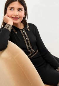 Czarna sukienka z prążkowanego materiału z kołnierzykiem i akcentami w leopardzim wzorze. Posiada dekoracyjne guziki we frontowym zapięciu. Miękka tekstura na beżowym krześle.