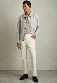Chaqueta bomber a cuadros grises con botones negros, combinada con pantalones color crema y zapatos de ante marrón, en un fondo apagado.