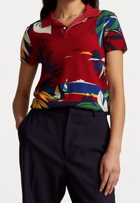 Kvinna i röd kortärmad polo med färgglada tropiska och segelbåtsmönster instoppad i mörka byxor, med händerna i fickorna.