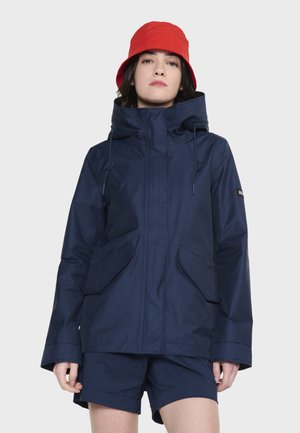Chaqueta impermeable azul marino con cuello alto, cordones ajustables y dos bolsillos frontales. Combinada con pantalones cortos azul marino y un sombrero de cubo rojo.
