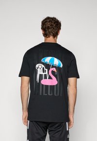 Sort t-shirt med et grafisk tryk af en pink flamingo, en hvid stol og en blå- og hvidstribet parasol på bagsiden. Blødt stof.