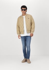 Chaqueta bomber acolchada beige con botones a presión, llevada sobre una camiseta blanca, combinada con jeans azules y zapatos de color claro. Gafas de sol negras.