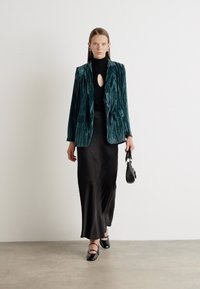 Teal velvet blazer with vertical stripes, black mock neck top, flowing black skirt, and black shoes with straps. Compact black bag.
