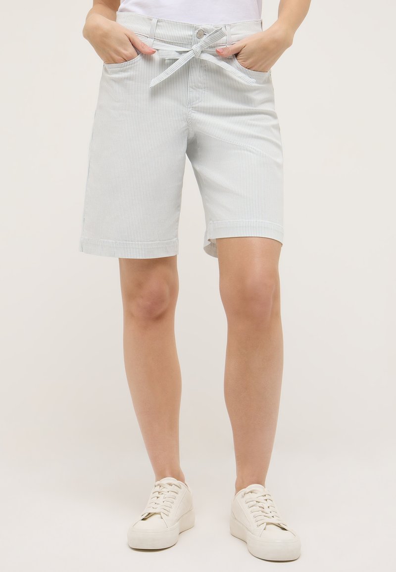 Femme portant un short Bermuda gris clair à fines rayures avec ceinture nouée à l'avant et baskets blanches, debout les mains dans les poches.