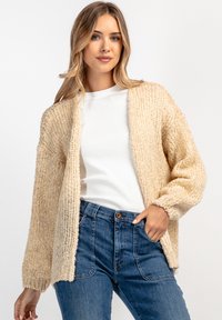 Beżowy sweter dzianinowy o luźnym kroju, teksturowanej tkaninie, z otwartym przodem i długimi rękawami, noszony na białej koszuli i niebieskich dżinsach.