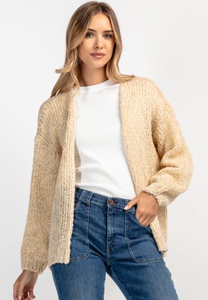 Beżowy sweter dzianinowy o luźnym kroju, teksturowanej tkaninie, z otwartym przodem i długimi rękawami, noszony na białej koszuli i niebieskich dżinsach.