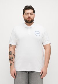 Cămașă polo albă din bumbac, cu guler, mâneci scurte și un logo circular albastru pe pieptul stâng. Textură simplă, croi clasic.