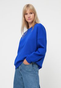 Hanorac albastru dintr-un material din bumbac amestecat, cu o croială lejeră, decolteu rotund și manșete din tricot, asociat cu blugi decolorati.
