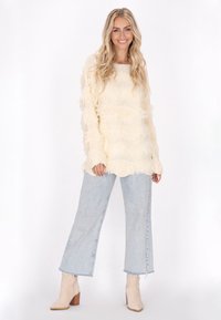 Maglione crema con frange e texture a maglia orizzontale, abbinato a jeans a gamba larga in lavaggio chiaro e stivaletti beige con tacco a blocco.