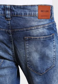 Jeans de mezclilla azul con un acabado desgastado y texturizado. Presentan un parche de cuero marrón etiquetado "ONLY & SONS" en la parte posterior de la cinturilla y dos bolsillos traseros.