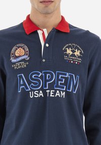 Tengeri kék hosszúujjú póló piros gallérral. Fehér és kék grafikákkal rendelkezik: "ASPEN USA TEAM" és több hímzett logó. Puha pamut anyag.