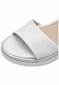 Valkoinen liukas sandaali, jossa on leveä, sileä nahkahihna ja teksturoitu beige pohja. Sandaalissa on hopeinen yksityiskohta pohjassa.