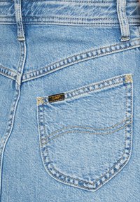 Ljusblå denimjeans har en liten bakficka med en svart "Lee"-etikett. Tyget har en texturerad yta med gula sömnadsdetaljer.