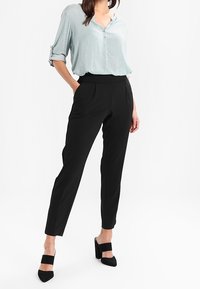 Blouse bleu clair à motifs avec manches retroussées et fermeture à boutons, associée à un pantalon noir ajusté et des chaussures à talons hauts noires.