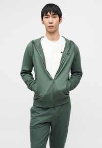 Roheline lukuga kapuutsiga pusa, valmistatud pehmest kangast, millel on küljetaskud, kantud valge T-särgi peal. Liibuv siluett, minimaalne brändingudetail.