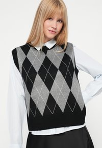 Fekete és szürke argyle kötött mellény bordás szegéllyel, egy világoskék galléros ing felett viselve. Gyémánt mintázattal és fehér varrási részletekkel rendelkezik.