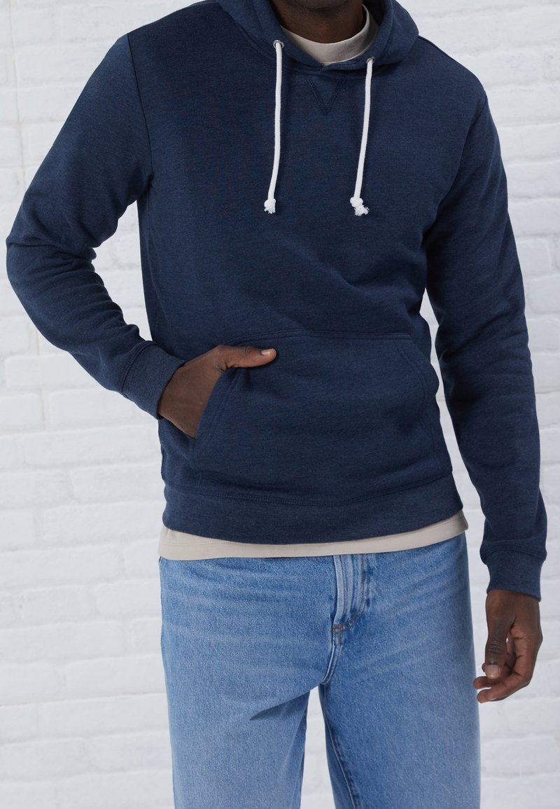 Mörkblå fleecehoodie med kenguruficka, vita dragsnören och ribbade manschetter, lager på en ljusfärgad skjorta och blå jeans.