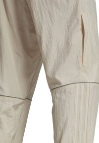 Light beige athletic pants with a textured finish, featuring a side pocket and subtle gray piping along the seams and pleats.