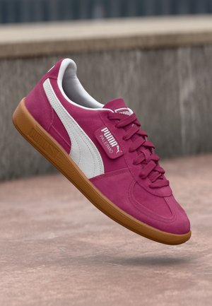 Zapatilla Puma Palermo de color rosa brillante con raya blanca y suela de goma, mostrada flotando sobre una superficie de cemento.