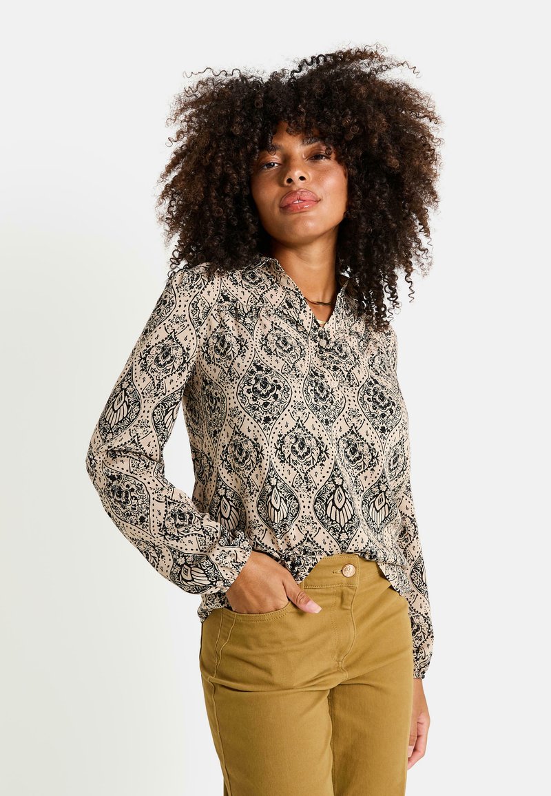 Blusa de manga larga con cuello en una tela beige, que presenta un patrón floral y de paisley en negro. Combinada con pantalones caqui, con un diseño ajustado.