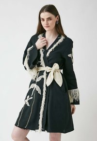 Svart cape med fransdetaljer och ett bälte; pryds av beige broderi av växter. Långa ärmar och en texturerad yta, knälång.