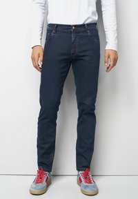 Jean en denim bleu foncé avec une coupe slim, cinq poches et des coutures discrètes. Porté avec des baskets bleu clair à lacets rouges.