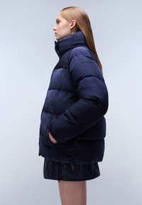 Nafukovací bunda v námořnické modři, s prošívanými částmi a vysokým límcem. Materiál vykazuje ribbed strukturu. Obsahuje boční kapsy a nášivku značky.