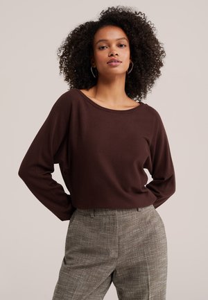 Femme aux cheveux bouclés portant un haut à manches longues marron foncé et un pantalon à carreaux gris, debout, les mains dans le dos.