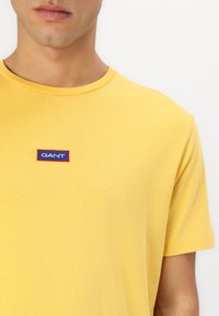 Tricou din bumbac galben, cu gât rotund; prezintă un mic logo brodat "GANT" în culorile albastru și roșu pe piept. Textură moale și design fără cusături.