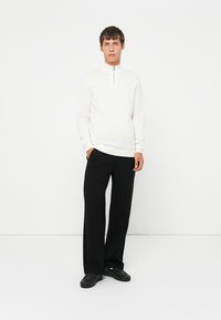 Weißes Langarmoberteil mit Viertelreißverschluss und gerippten Bündchen, kombiniert mit schwarzen weiten Hosen und schwarzen Schuhen. Minimalistisches Design.