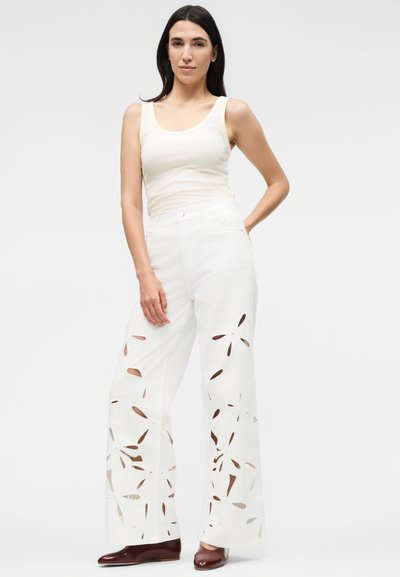 Γυναίκα με μακριά σκούρα μαλλιά φοράει αμάνικη μπεζ μπλούζα και λευκό παντελόνι με φαρδιά πόδια και λουλουδάτα κοψίματα, συνδυασμένα με σκούρα καφέ παπούτσια.