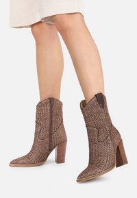 Botas trenzadas marrones hasta la mitad de la pantorrilla con tacones de madera gruesos y cremalleras laterales, llevadas con pantalones cortos de color beige claro.