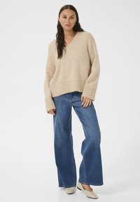 Maglione oversized beige a scollo a V con orlo a costine, abbinato a jeans blu a gamba larga e scarpe slip-on beige. Tessuto morbido e vestibilità casual.