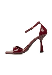 Sandalia de tacón alto de charol rojo con tira en el tobillo, punta abierta y un elegante tacón de aguja. Textura suave y brillante con un diseño minimalista.