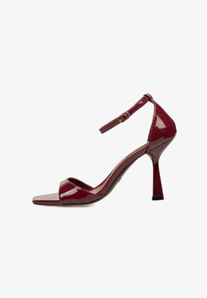 Sandalia de tacón alto de charol rojo con tira en el tobillo, punta abierta y un elegante tacón de aguja. Textura suave y brillante con un diseño minimalista.