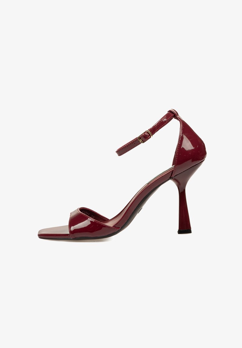 Sandalia de tacón alto de charol rojo con tira en el tobillo, punta abierta y un elegante tacón de aguja. Textura suave y brillante con un diseño minimalista.