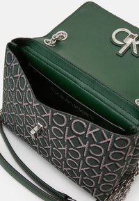 Grøn lædertaske med monogrammet stof og sølvfarvet kædetras, åbent låg der viser Calvin Klein-logoet indeni.
