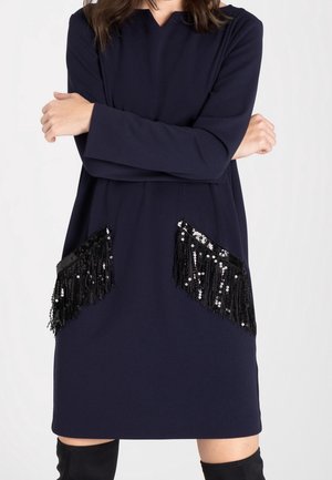 Woman wearing a navy long-sleeve dress with black sequin fringe pockets and crossed arms against a plain background.