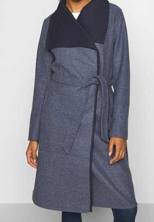 Femme portant un manteau en laine gris ceinturé, longueur genoux, avec un large col châle bleu marine foncé.