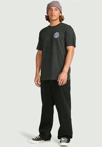 T-shirt preta de manga curta com decote redondo e logótipo frontal, acompanhada de calças pretas largas e sapatilhas pretas. Gorro de malha roxa utilizado.